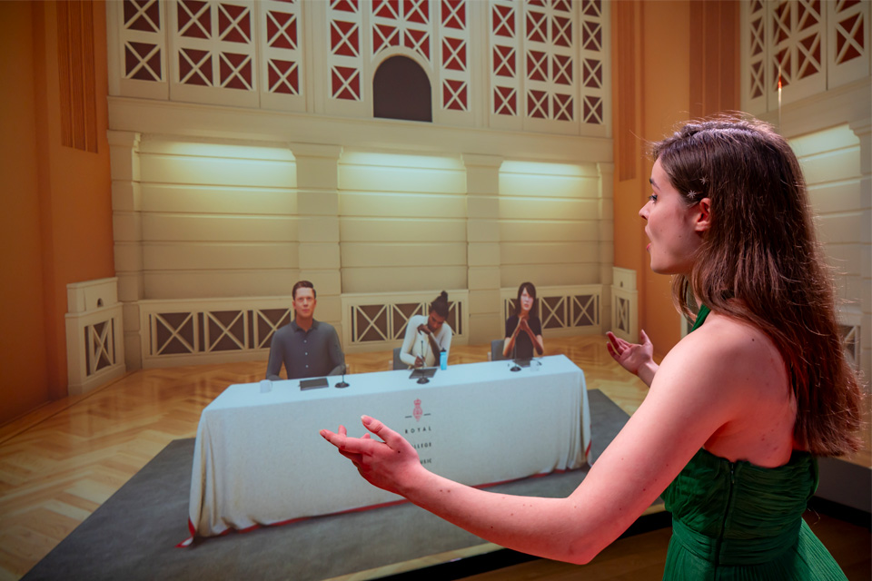 A student wearing a green dress, singing in front of a fake audience in a simulator.